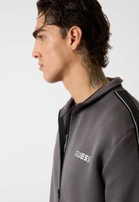 Felpa grigia con zip, caratterizzata da un logo, un accento nero con motivo laterale e una texture morbida. Design frontale con cappuccio.