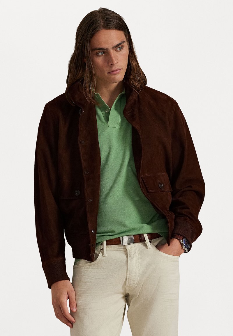 Bruin suède jack met een afneembare capuchon, twee voorzakken, gedragen over een groene polo en lichte broek. Casual ontwerpkenmerken.