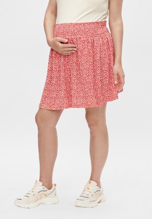 Femme portant une jupe florale rouge et blanche, un haut blanc et des baskets blanches, une main reposant sur la taille de la jupe.