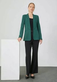 Blazer teal com um corte ajustado, mangas longas e design aberto à frente, combinado com calças largas pretas e sapatos pretos. Textura de tecido suave.