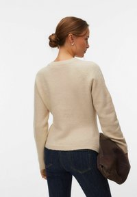 Femme aux cheveux bruns attachés en chignon, portant un pull beige et un jean foncé, tenant une veste en daim brun foncé pliée dans sa main gauche.
