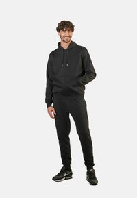 Svart hoodie med en känguruficka, dragsnören och matchande joggers med ribbade ärmslut, tillverkad i ett mjukt tyg. Svarta sneakers fulländar outfiten.