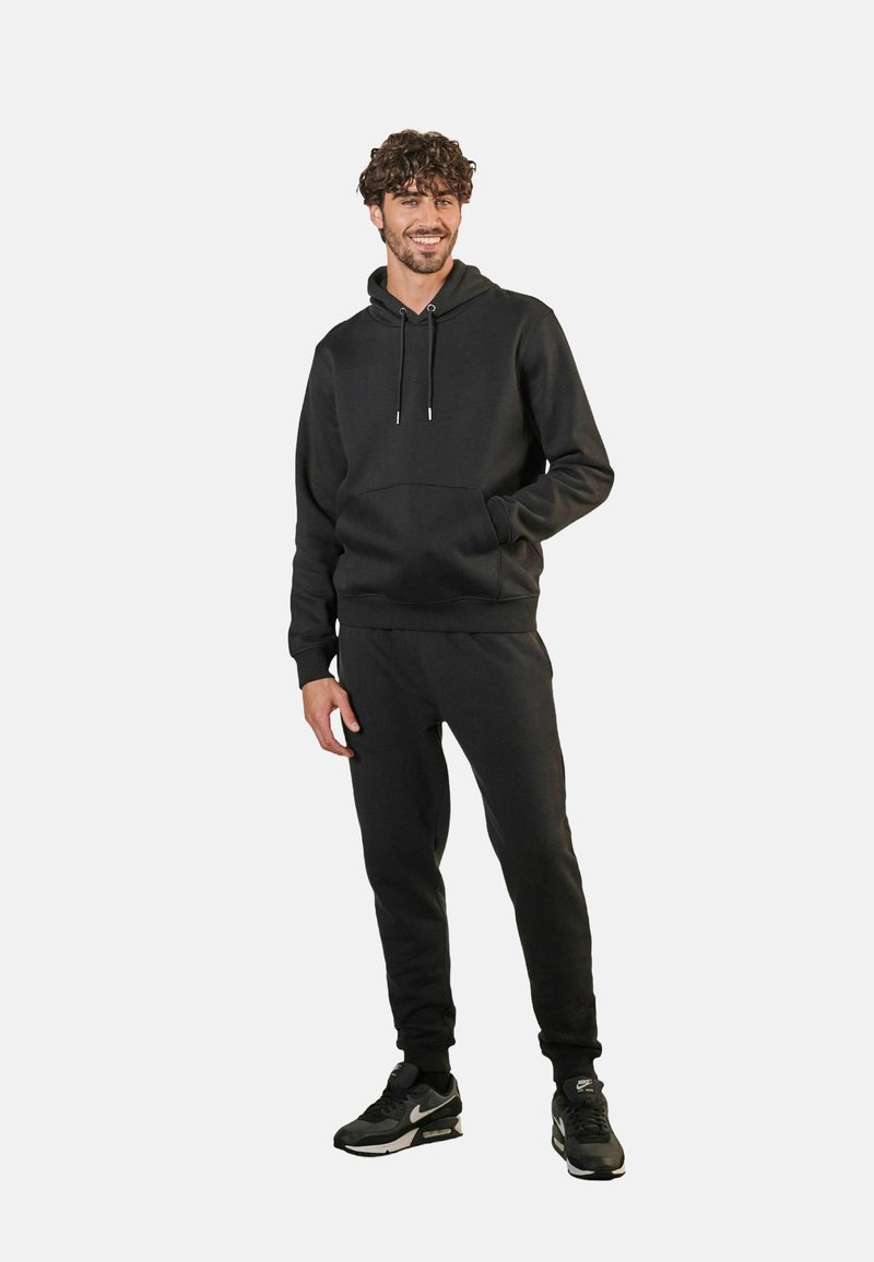 Svart hoodie med en känguruficka, dragsnören och matchande joggers med ribbade ärmslut, tillverkad i ett mjukt tyg. Svarta sneakers fulländar outfiten.