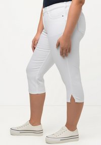 Los pantalones capri blancos presentan un diseño ajustado con aberturas en los laterales del dobladillo. Se combinan con zapatillas blancas y una camiseta azul marino.