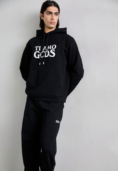 La sudadera negra presenta "TI AMO GCODS" en texto blanco, combinada con pantalones de chándal negros que exhiben un pequeño logo blanco. Tejido suave, corte informal.
