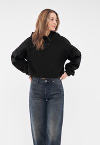 Felpa nera corta con cappuccio regolabile e maniche ampie, caratterizzata da un logo discreto. Abbinata a jeans in denim scuro a vita alta.