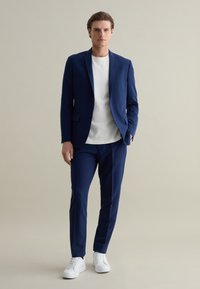 Completo blu navy con giacca aderente e pantaloni, abbinato a una maglietta bianca a girocollo e sneakers bianche. Tessuto liscio, design minimale.
