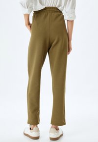 Olijfgroene geribde broek met een elastische tailleband, slim fit, en zijsplits aan de zoom, gecombineerd met witte sneakers met lichte accenten.