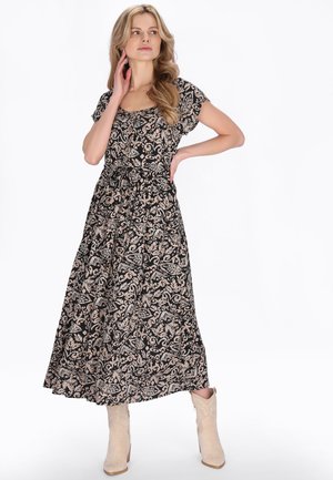 Vrouw draagt een zwarte maxi-jurk met beige bloemachtige patronen, korte mouwen, een gestrikte taille en beige enkellaarzen, poseert met hand op heup.