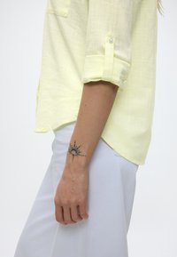 Άτομο που φορεί κίτρινο πουκάμισο με γυρισμένα μανίκια και λευκό παντελόνι, με ένα μαύρο τατουάζ στον καρπό σε σχήμα ήλιου στο αριστερό του χέρι.