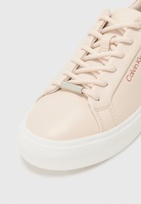 Sneaker in pelle beige con suola in gomma bianca, caratterizzata da lacci piatti e un accento argento con il marchio "Calvin Klein" sul lato.
