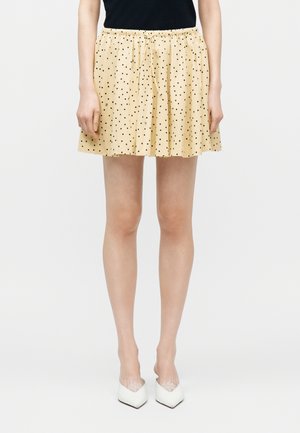 Žena v béžovej mini sukni s čiernymi bodkami, čiernom topu a bielych vysokých opätkoch s hrotmi stojaca pred jednoduchým pozadím.