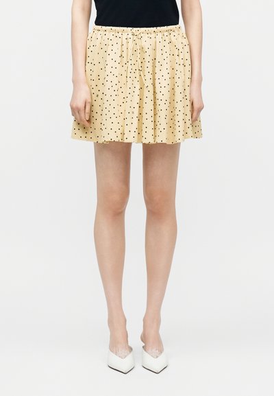 Femme portant une mini-jupe beige à pois noirs, un haut noir et des chaussures blanches à talons aiguilles pointues, debout devant un fond uni.