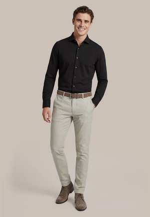 Lächelnder Mann, der ein schwarzes Hemd, beige Hose mit braunem Gürtel und braune Schnürschuhe trägt, steht mit einer Hand in der Tasche.