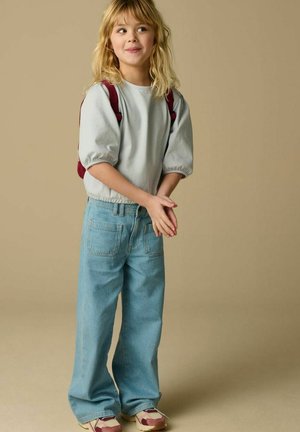 Jeune enfant aux cheveux blonds portant un t-shirt rayé, un jean bleu clair à jambes larges, des baskets et un sac à dos rouge, debout devant un fond beige uni.