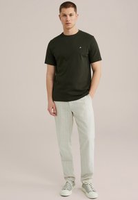 Temno zelena bombažna majica s krožnim ovratnikom in kratkimi rokavi, v kombinaciji s svetlimi, trakastimi hlačami sproščenega kroja in sivimi športnimi copati.