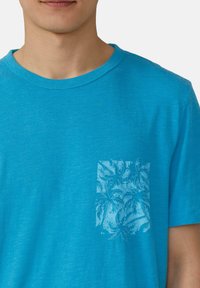 Turquoise katoenen T-shirt met een ronde hals en een borstzak met een lichtblauwe palmbladpatroon. Zachte textuur, casual ontwerp.