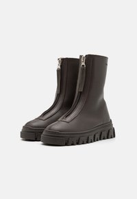 Bocanci din piele maro la gleznă, cu fermoare duble frontale, tălpi din cauciuc groase cu caneluri și detalii de cusătură proeminente. Design elegant și modern.