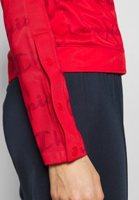 Gros plan sur le bras d'une personne portant une veste rouge à boutons-pression et un pantalon foncé, mettant en valeur les détails du poignet et de la manche de la veste.