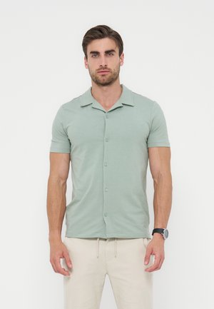 Mężczyzna ubrany w jasnozieloną koszulę z krótkim rękawem na guziki i beżowe spodnie, stojący na jednolitym białym tle.