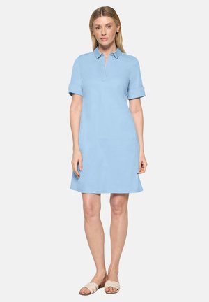 Frau steht und trägt ein hellblaues, kurzärmeliges Kleid mit Kragen und hochgekrempelten Ärmeln sowie beige flache Sandalen vor einem einfarbigen Hintergrund.