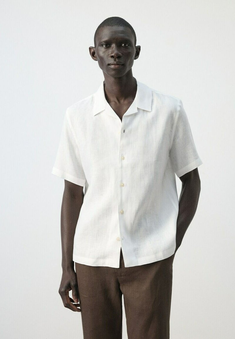 Homme à la peau foncée portant une chemise blanche à manches courtes et un pantalon marron, debout devant un fond clair uni.