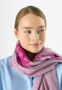 Lichtpaars sjaal met roze patroonaccenten, gemaakt van zachte stof, heeft franjes aan de randen, gewikkeld om de nek, gedragen over een blauwe shirt.