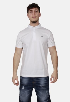 Uomo che indossa una polo bianca a maniche corte con bottoni e pantaloncini di jeans scuri slavati, in piedi davanti a uno sfondo semplice.