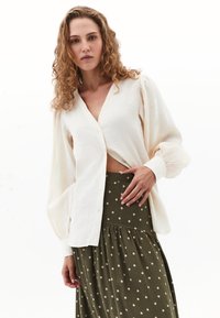 Crème knoopblouse met vloeiende mouwen, gecombineerd met een lange olijfgroene rok met witte stippen en een geraffineerde taille.