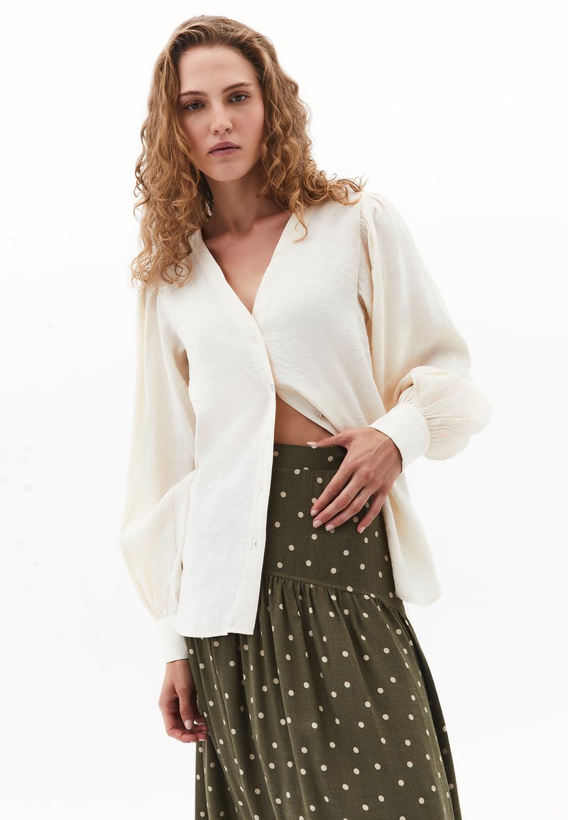 Crème knoopblouse met vloeiende mouwen, gecombineerd met een lange olijfgroene rok met witte stippen en een geraffineerde taille.