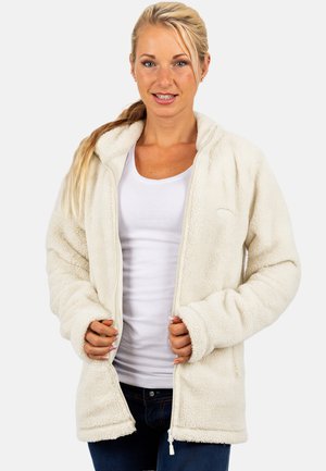 Jonge vrouw met blond haar die een wit fleecejack draagt over een wit overhemd en blauwe spijkerbroek, terwijl ze het ritsgedeelte van het jack vasthoudt.