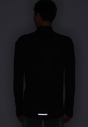 Mann trägt schwarzen Langarmshirt mit reflektierendem Streifen im unteren Rückenbereich, steht vor dunkelgrauem Hintergrund, mit dem Rücken zur Kamera.