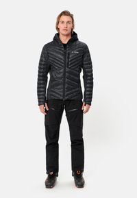 Giacca nera isolata con motivo a zigzag trapuntato, chiusura con zip e cappuccio. Abbinata a pantaloni neri e scarponi da sci con accenti arancioni.