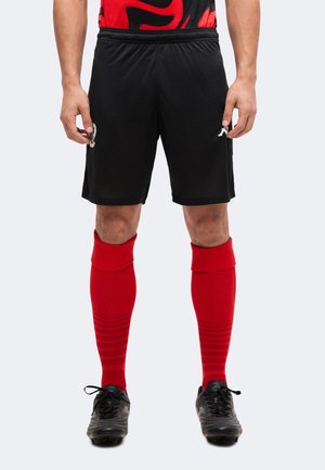 Man draagt zwarte sportshorts, rode kniekousen en zwarte voetbalschoenen, staand tegen een effen achtergrond.
