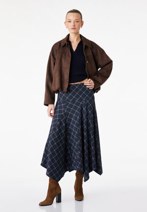 Vrouw staand, draagt een bruin suède jasje, zwart cropped topje, blauwe geruite asymmetrische rok en bruine laarzen met hak op witte achtergrond.