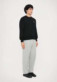 Felpa nera in cotone con polsini a costine, abbinata a pantaloni della tuta grigi chiari in cotone e sneakers nere su uno sfondo neutro.