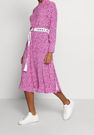 Persoon die loopt en een roze midi-jurk met bloemenpatroon en lange mouwen draagt, een witte Tommy Jeans-riem en witte sneakers, tegen een effen lichte achtergrond.