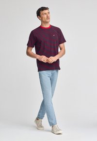 Kortärmad marinblå T-shirt med röda och blå ränder, med en röd halsringning och en liten broderad logotyp. Bärs med ljusblå jeans och sneakers.