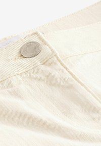 Beżowy denim o teksturowanej powierzchni, z srebrnym guzikiem w talii i delikatnym szwem wzdłuż szwów.