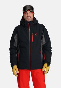 Black ski jacket with red accents, grey side panels, and hood. Worn with red pants and beige gloves. Ski goggles on the head.