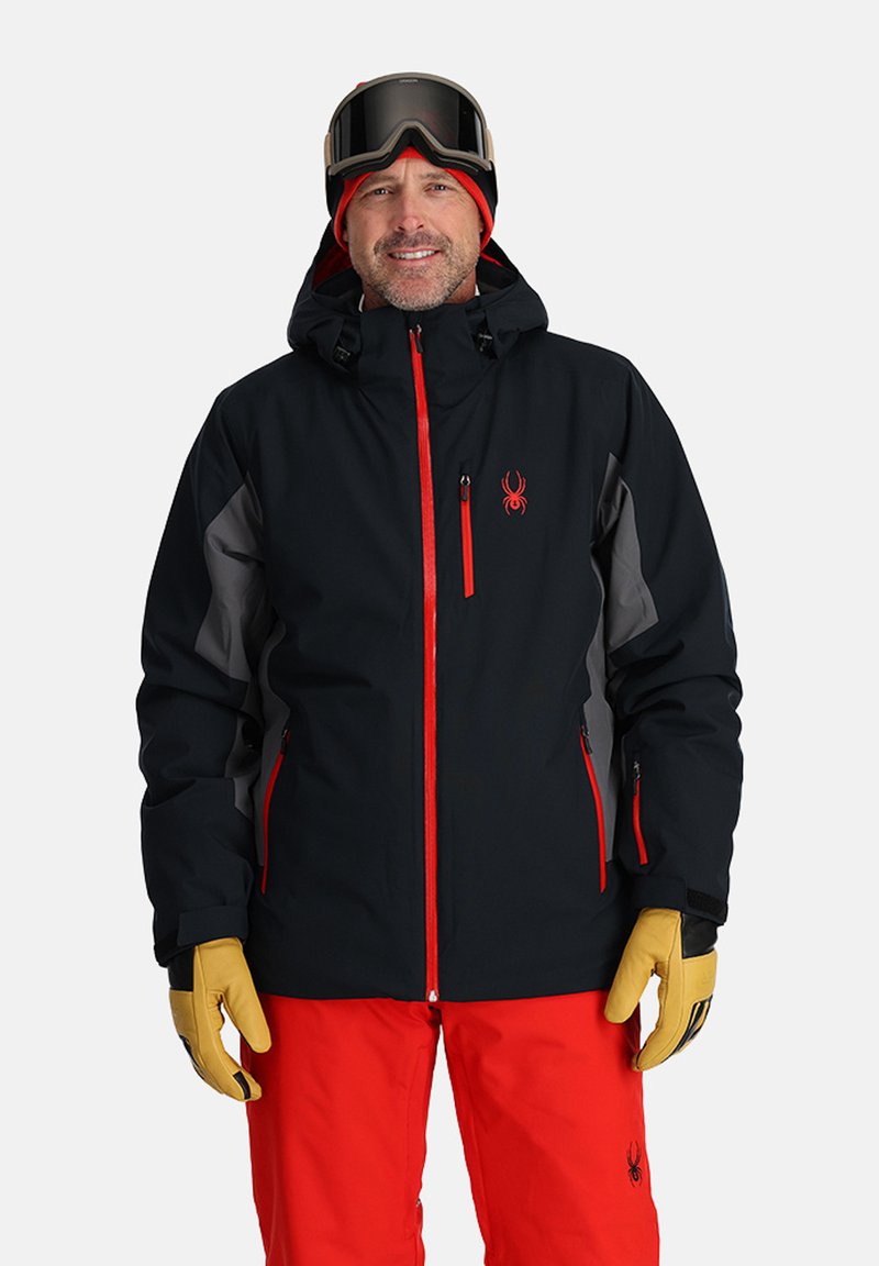 Black ski jacket with red accents, grey side panels, and hood. Worn with red pants and beige gloves. Ski goggles on the head.