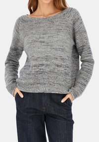 Kobieta ubrana w szary melanżowy sweter z długimi rękawami i ciemnoniebieskie dżinsy, ręce w kieszeniach, na jednolitym jasnym tle.