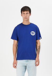 T-shirt in cotone blu con maniche corte, collo rotondo e logo rotondo che mostra "981 REPLAY" sul petto. Presenta una vestibilità rilassata.