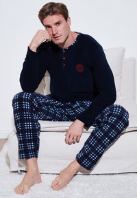 Marineblå langærmet top med ternet halsudskæring og røde knapdetaljer, parret med ternede fleece-pyjamasbukser i blå, rød og hvid.