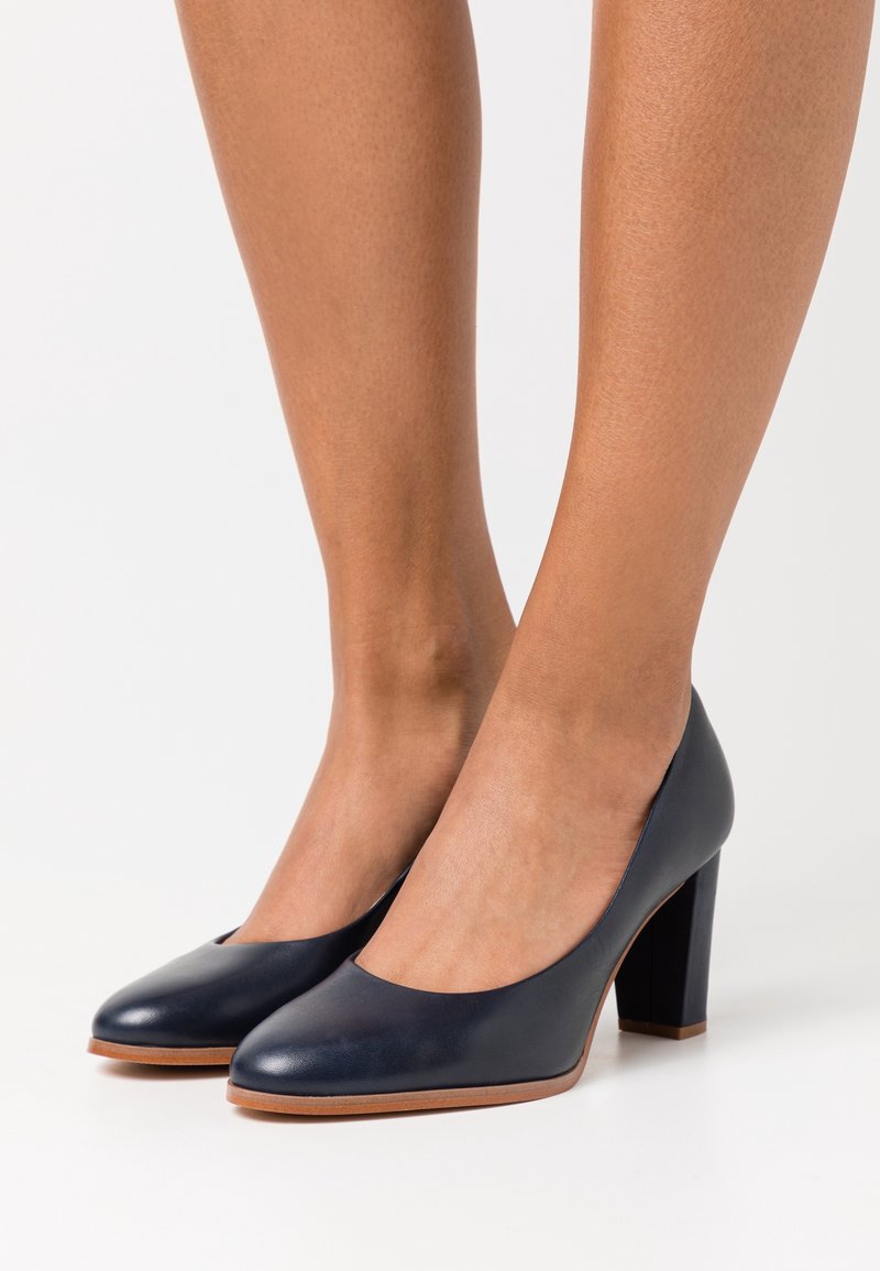 Des pieds portant des chaussures à talons hauts en cuir bleu marine avec des bouts ronds et des semelles beige, sur fond blanc.