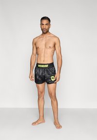 Melnas satīna šorti ar dzeltenu vidukļa joslu un logotipu, ar brīvu piegriezumu un gludu faktūru. Nav redzami raksti vai papildu akcenti.