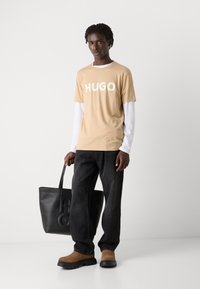 Šviesiai ruda T-marškiniai su balta "HUGO" logo, dėvimi ant baltų ilgomis rankovėmis marškinių, derinami su juodomis džinsais ir rudojo odos batais, laikantis juodą tvirtą maišą.