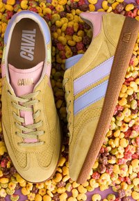 Baskets en daim jaune avec des rayures violet clair et des accents roses, placées sur un fond multicolore de céréales. Marque "CAVAL" visible à l'intérieur.