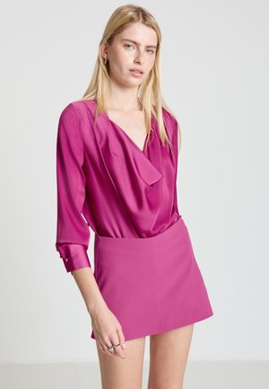 Femme aux cheveux blonds raides portant un chemisier en satin magenta à manches longues et une jupe courte assortie, debout devant un fond uni.