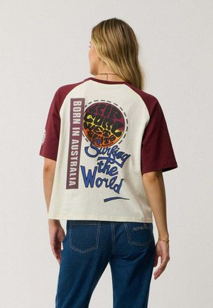 Vrouw met lang blond haar die een crème- en bordeauxkleurig kortemouw T-shirt met surfertekst draagt en een blauwe spijkerbroek, van achteren gezien.